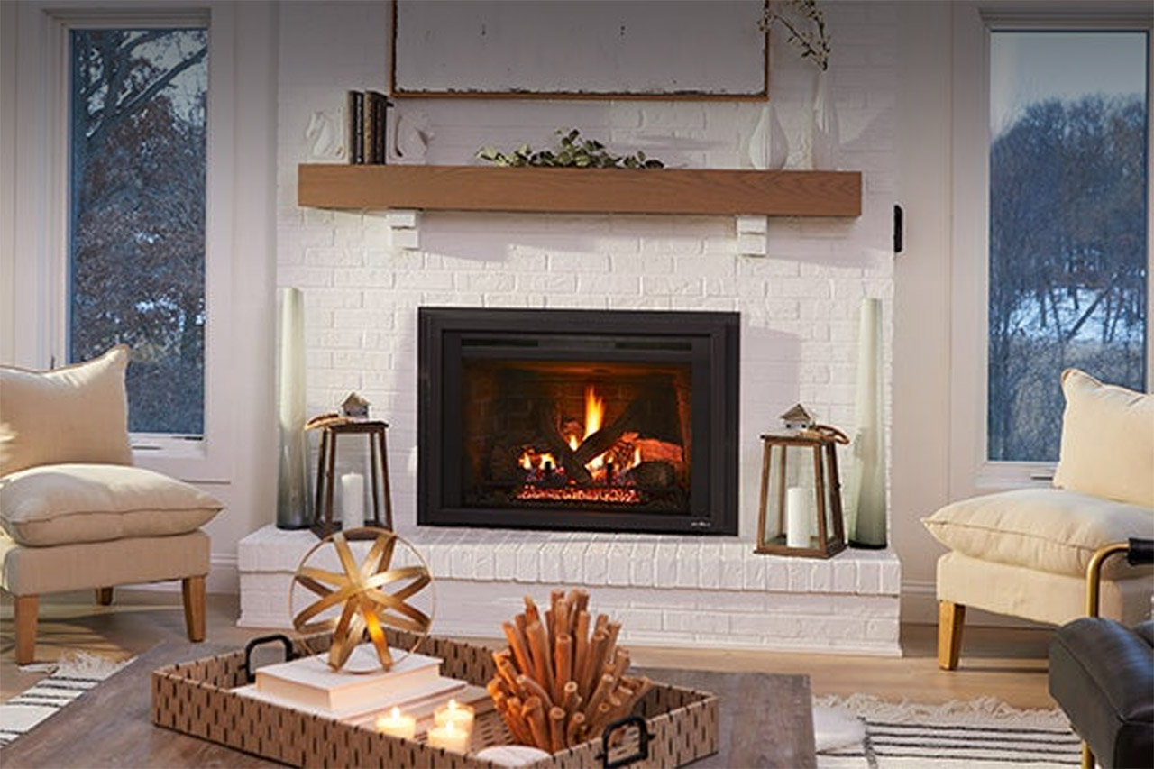 Cozy living room with a lit fireplace insert, wooden mantel, and white brick surround. Two lanterns and a decorative wooden star on a coffee table, with a snowy view outside the window.