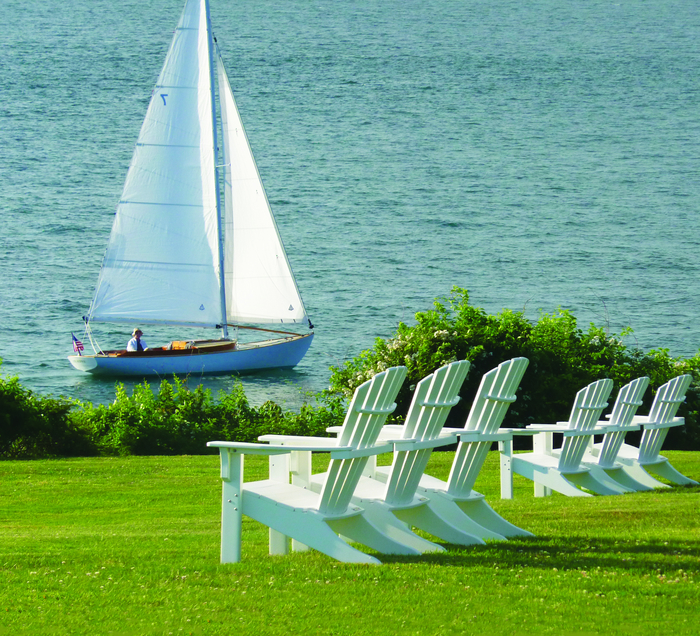 A sailboat with two passengers glides on blue water beside a grassy shore lined with white adirondack chairs and a fire pit facing the lake.