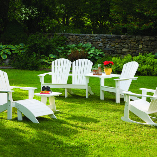 Four white adirondack chairs arranged in a semi-circle around a fire pit on a lush green lawn, with a small table in the center, surrounded by trees and a stone wall.