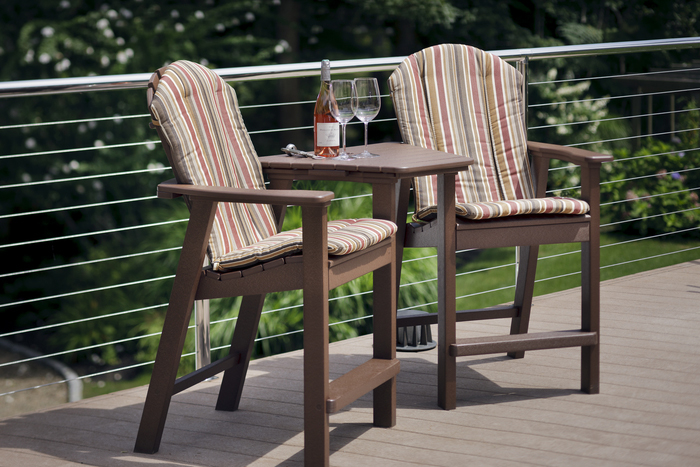 Two striped cushioned patio chairs with a small table between them on an outdoor deck, near a fire pit. The table holds a bottle of wine, two glasses, and a small plate with snacks,