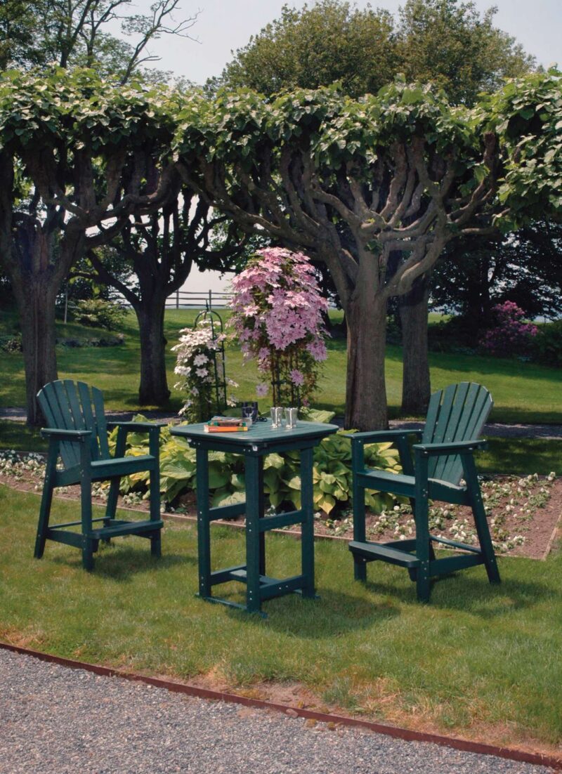 A serene outdoor dining setup with a green table and chairs under trees with lush foliage and pink flowering shrubs, overlooking a well-manicured lawn, featuring an elegant fireplace.