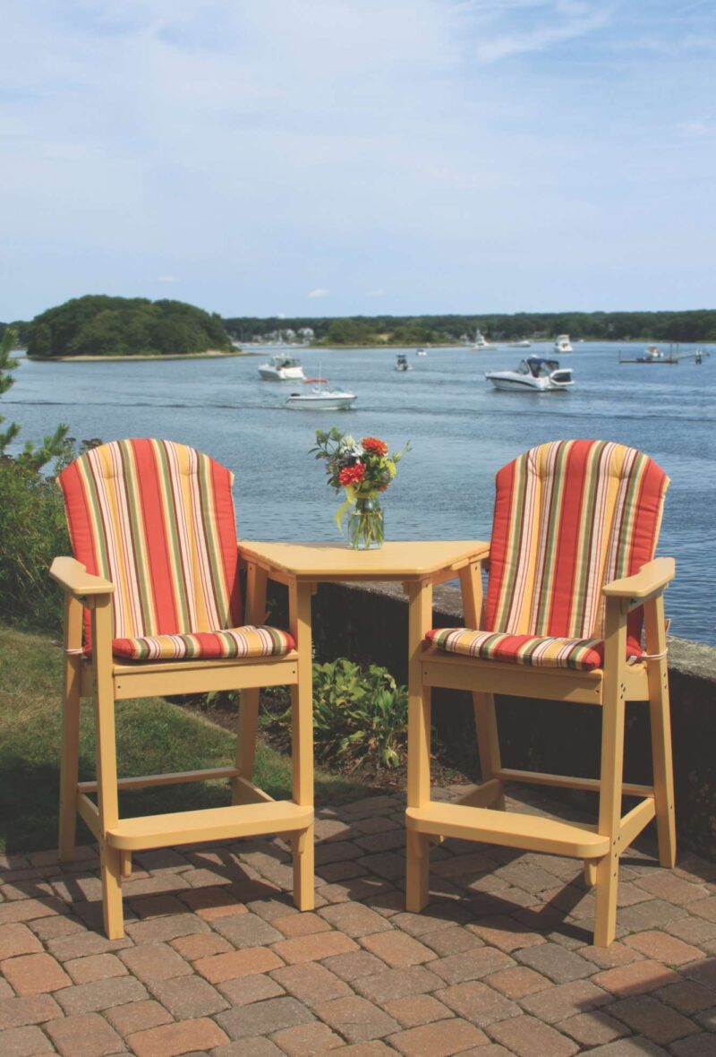 Two striped deck chairs facing a calm bay with boats. Between the chairs is a small wooden table with a vase of flowers and a fire pit nearby. The setting is peaceful and scenic.