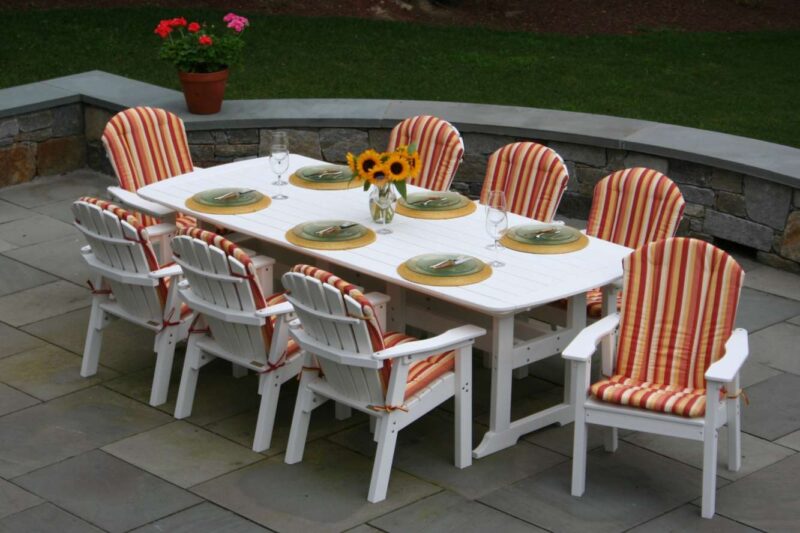 An outdoor dining area with a white table set for six, featuring stripe-cushioned chairs, plates, wine glasses, and a vase with sunflowers, all on a stone patio beside a fire pit