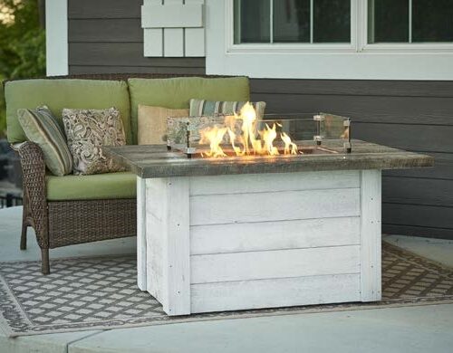 Outdoor patio with a lit fireplace table surrounded by a wicker loveseat and two chairs with green cushions, situated on a concrete pavement near a house.
