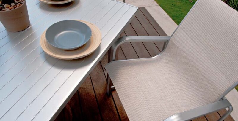 A modern outdoor dining setup featuring a light grey table with a stack of blue and beige plates on top, next to a silver-framed chair with beige fabric, set on a wooden deck near a cozy