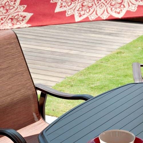 Outdoor patio setup with two brown wicker chairs around a small gray table, displaying a white vase and a cup, against a vibrant, patterned red throw on a garden fence near a cozy fireplace.