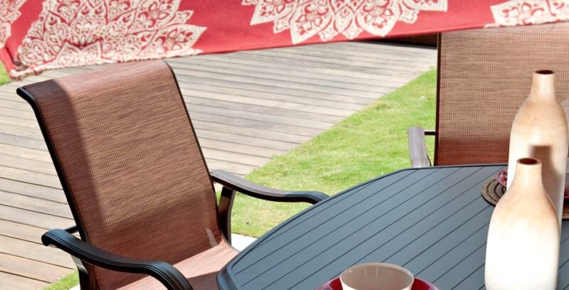 Outdoor patio setup with two brown wicker chairs around a small gray table, displaying a white vase and a cup, against a vibrant, patterned red throw on a garden fence near a cozy fireplace.