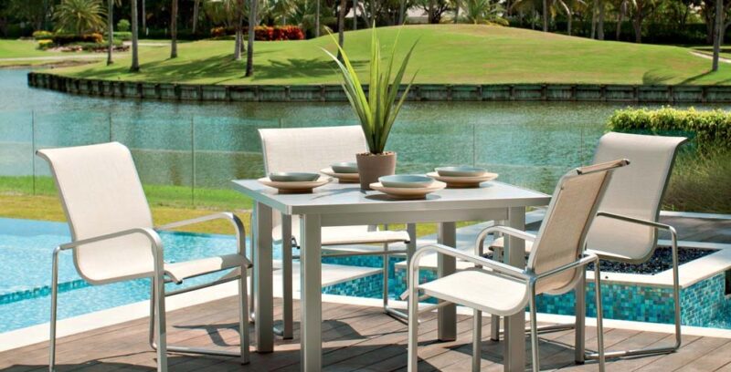 Outdoor dining area featuring a modern table set with four chairs and a cozy fireplace insert, overlooking a tranquil pool and lush green golf course.