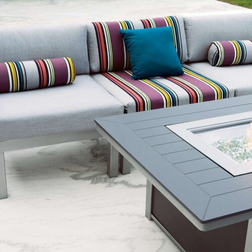 Outdoor patio with a modern gray sectional sofa adorned with colorful striped cushions and a central rectangular fireplace table, set on a smooth marble floor.