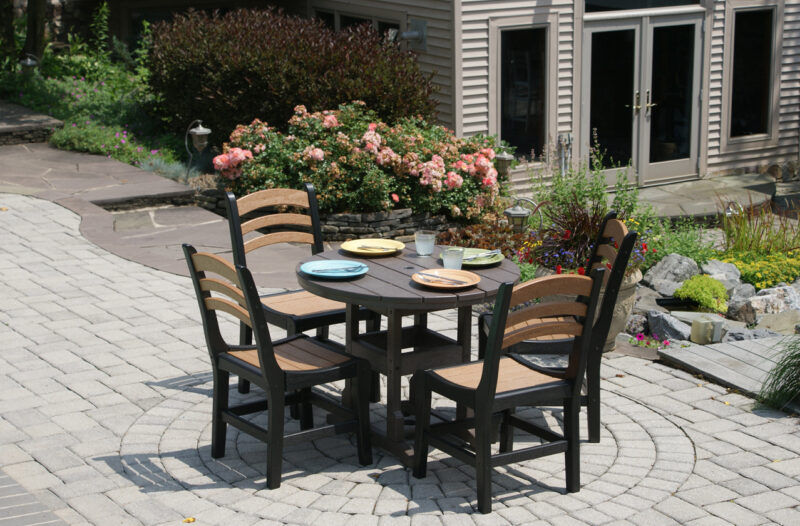 Outdoor patio dining area with a circular table set with colorful plates, surrounded by four chairs, next to blooming rose bushes and a house, featuring an inviting fire pit.
