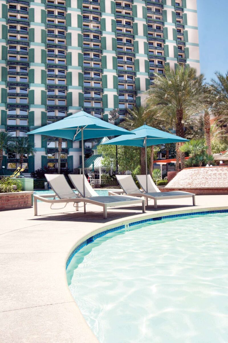Two lounge chairs under a blue umbrella by a poolside, with a tall hotel building and palm trees in the background, featuring an outdoor fireplace on a sunny day.