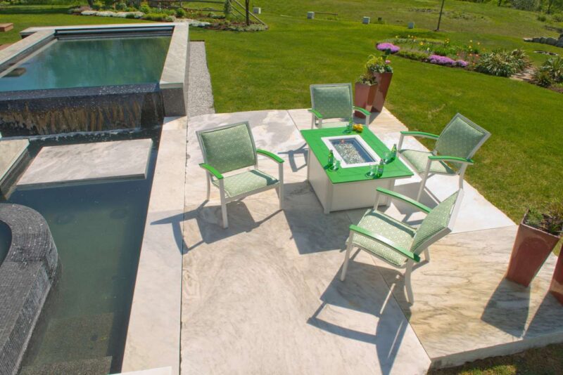 A sunny outdoor scene with a modern swimming pool adjacent to a patio area featuring four green chairs around a square table and an inserted pizza oven, set against a backdrop of lush green grass and colorful flowers.