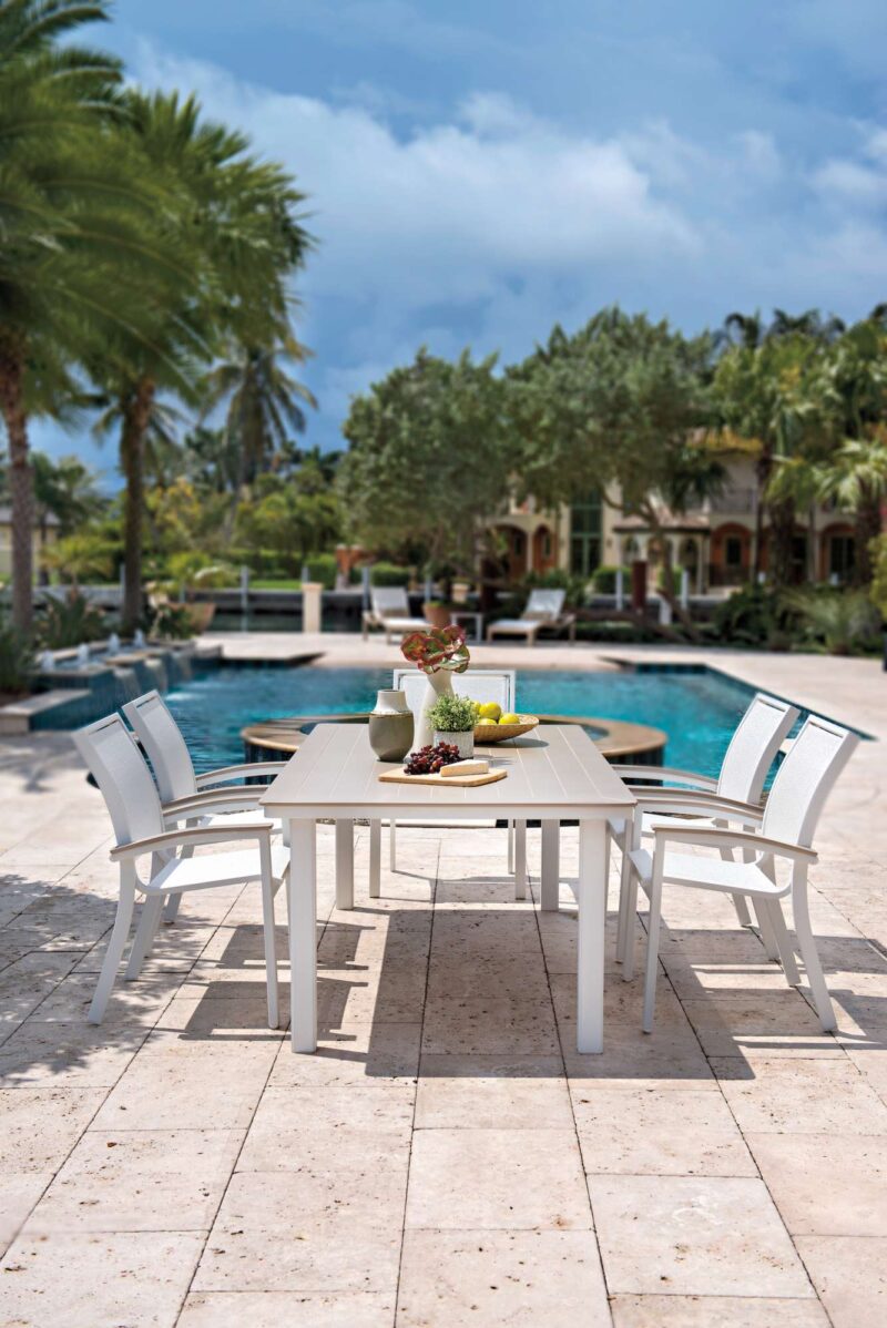 Outdoor dining setup by a pool, featuring a white table with chairs, various dishes including a nearby grill, and a luxurious home surrounded by palm trees under a blue sky.