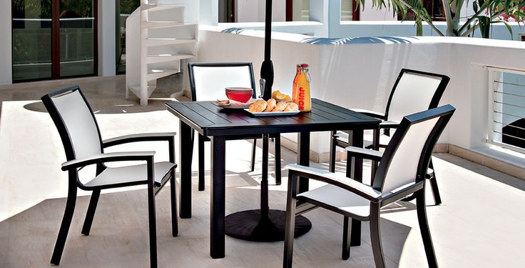 A modern outdoor dining area featuring a black table and four chairs, set with bread, olives, and a bottle of wine on a sunny patio with a fireplace.