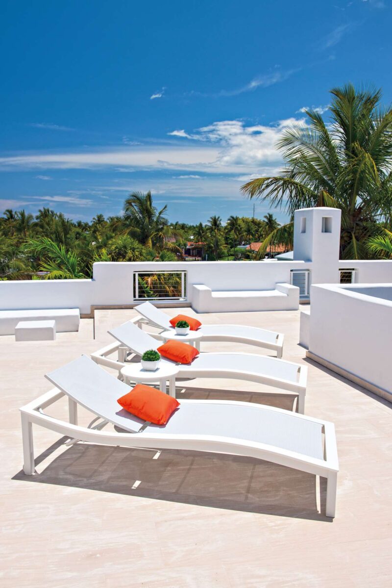 A rooftop terrace with white lounge chairs adorned with orange cushions, surrounded by lush greenery under a blue sky, features a sleek grill.