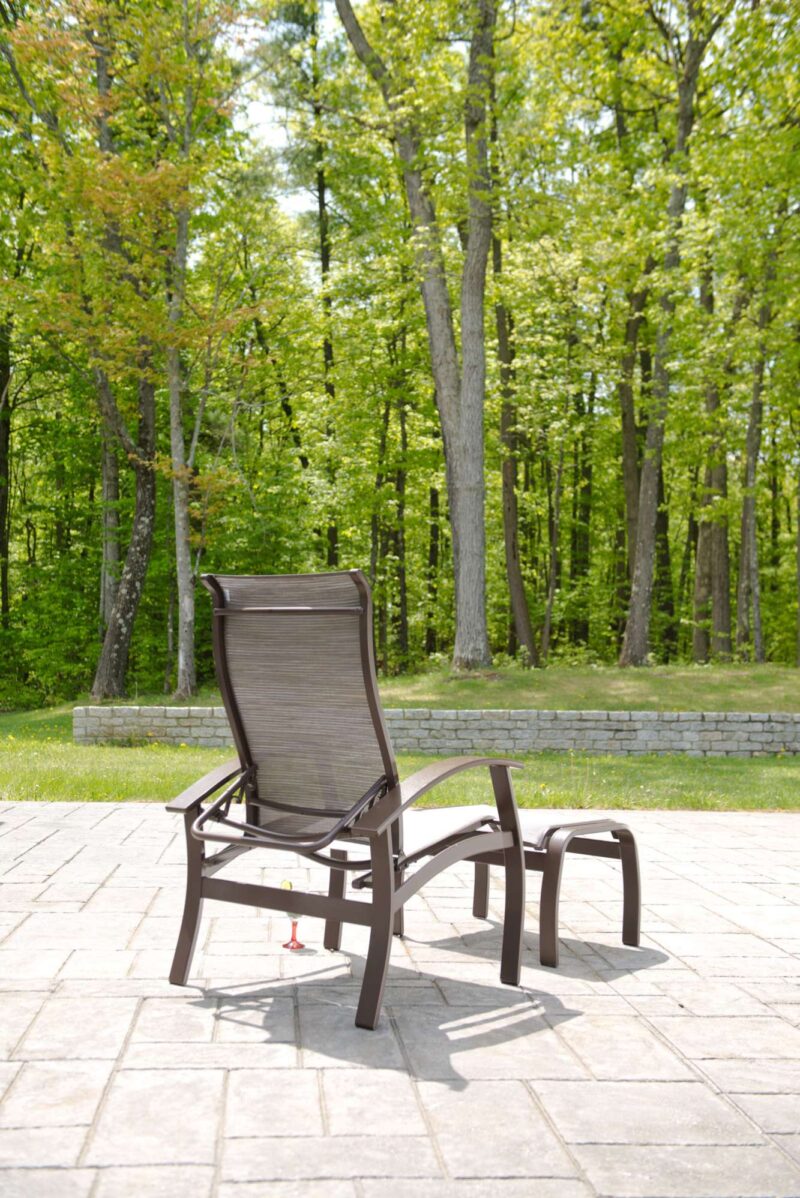 An empty reclining deck chair made of dark metal with a fabric seat and backrest, positioned on a paved patio surrounded by lush green trees and a fireplace under a bright, clear sky.