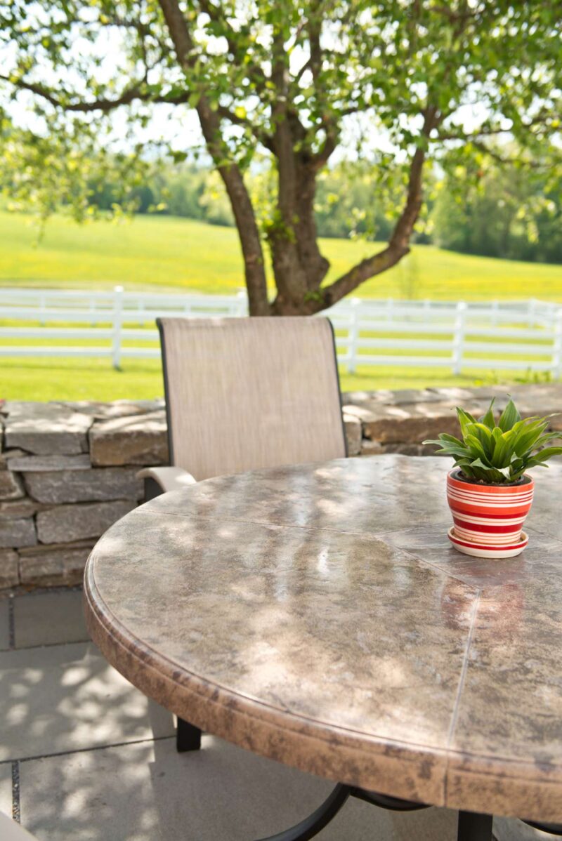 A serene outdoor setting featuring a round stone table with a potted plant, a single chair, and a view of a lush green landscape enclosed by a white fence under a tree, complemented by an