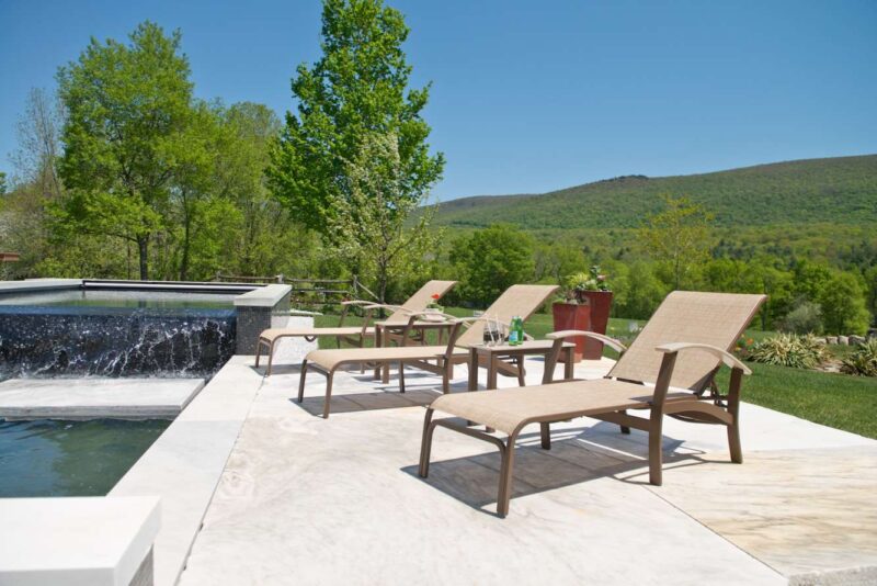 Outdoor patio with lounge chairs and a table beside an infinity pool overlooking a lush mountain landscape, under a clear blue sky, featuring a grill.