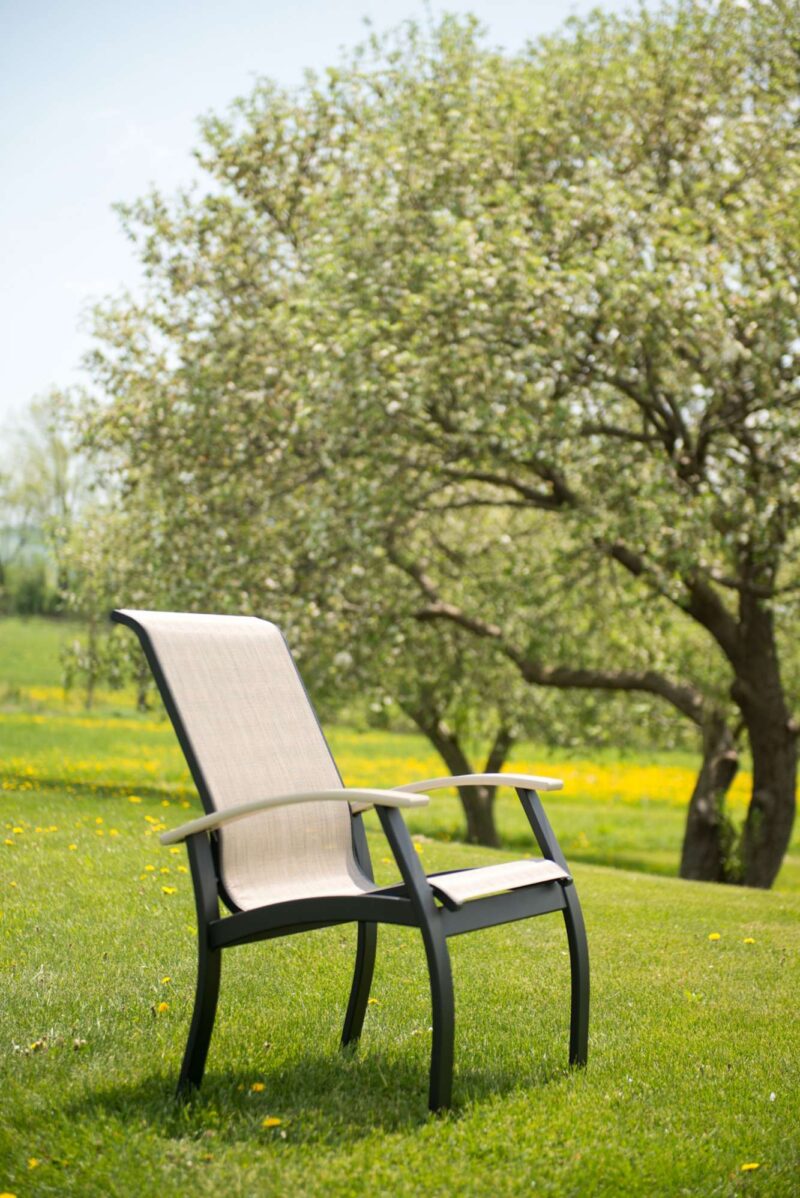 A beige outdoor chair stands alone in a lush green field with dandelions, near a fire pit, under blossoming apple trees with a soft focus background.