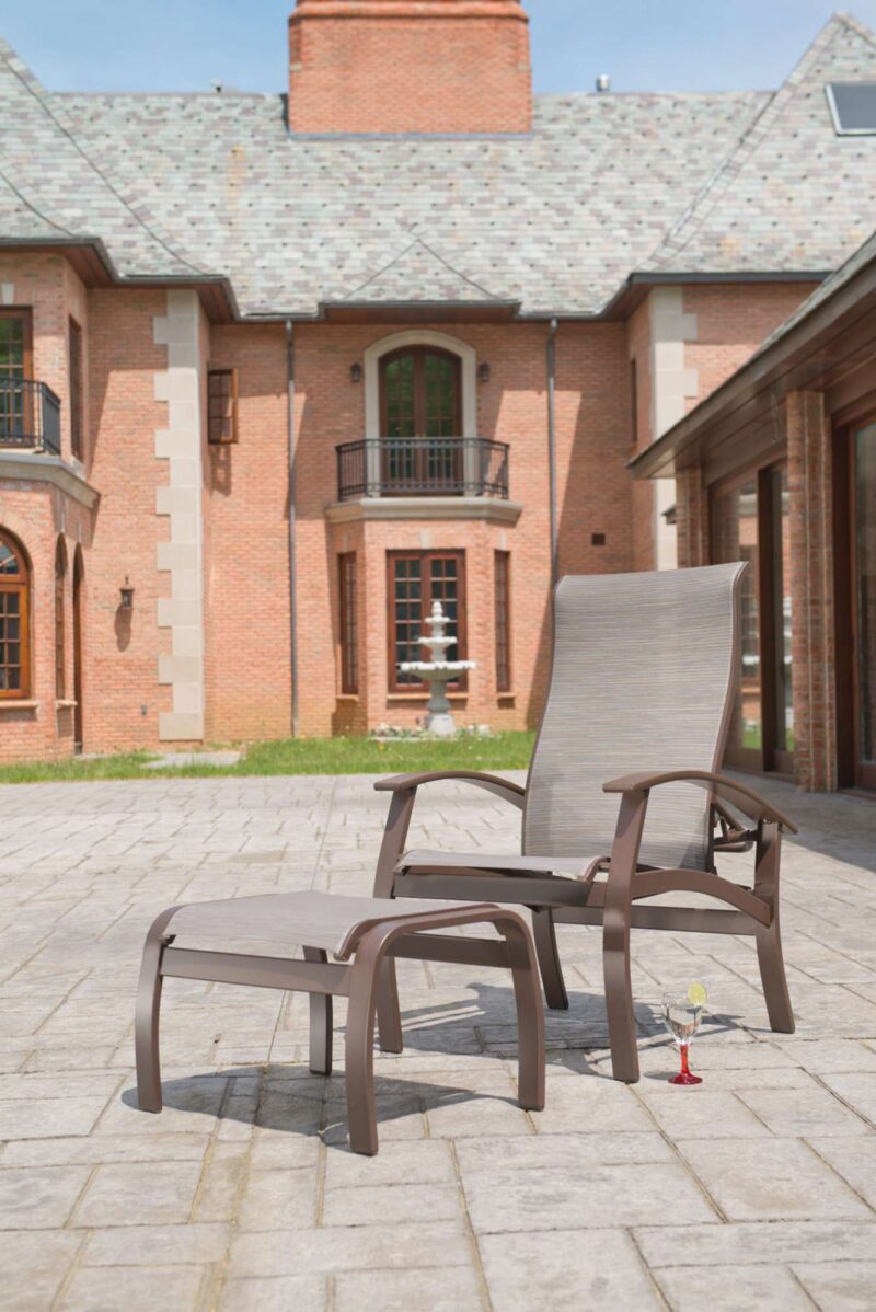 A reclining lounge chair and a small side table with a cocktail on a sunlit patio, featuring an elegant brick house with a fireplace in the background.