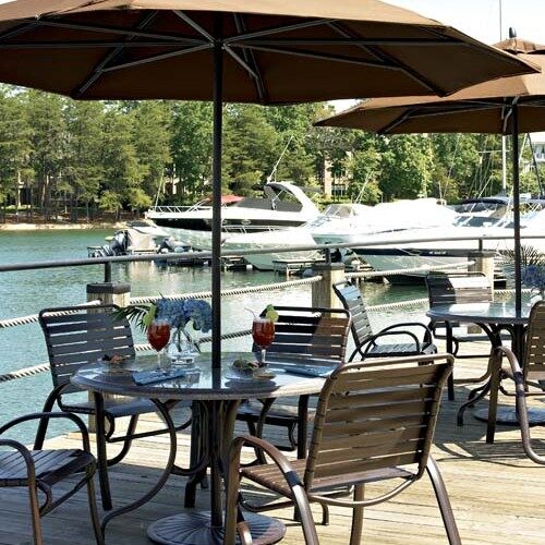 Outdoor lakeside dining area with several tables under umbrellas, overlooking a dock with moored boats. A pizza oven is visible in the background, and drinks are on a table in the foreground.