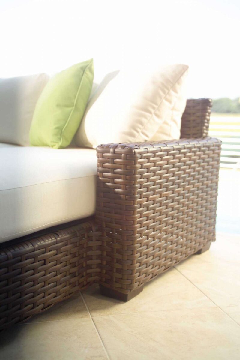 A close-up of a wicker sofa with white cushions and green pillows, placed outdoors near a fire pit, with a soft focus background suggesting a sunny, pleasant setting.