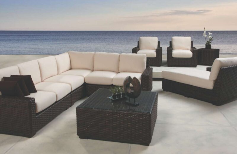 An outdoor patio setup with a sectional and individual rattan chairs, white cushions, and a center table with an inserted fire pit, overlooking a calm sea during twilight.