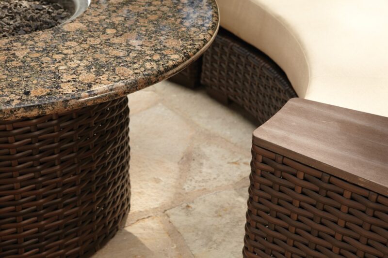 Close-up of a patio corner featuring a curved wicker sofa with light cushions, a round wicker table with an insert granite top, and a matching wicker footrest, all set on stone tiles