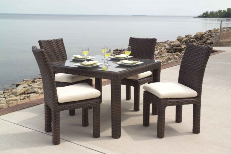 Outdoor dining set with four brown wicker chairs and a matching table, set with dishes and glasses, overlooking a calm lake featuring a fire pit.