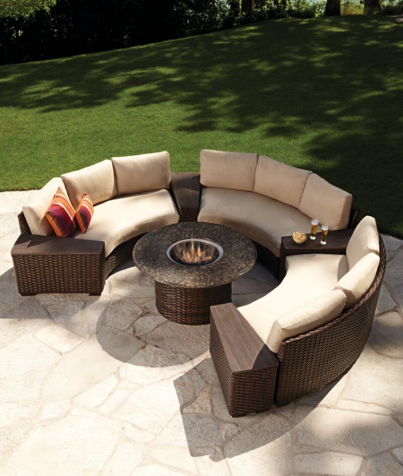 Outdoor patio furniture set with a circular design, including a central fireplace, surrounded by curved sofas with cushions, in a lush garden setting.