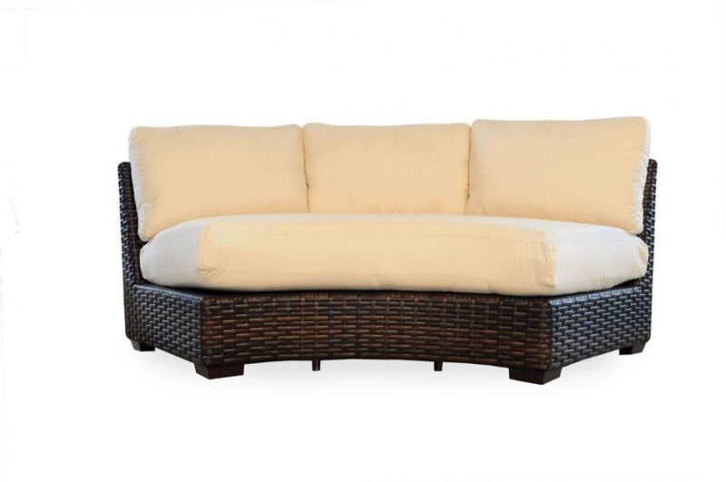 A curved wicker sofa with a dark brown frame and three large, beige cushions beside a fireplace on a white background.