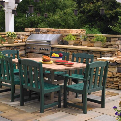 Outdoor patio setting featuring a stainless steel grill, a dining table with six green chairs, and surrounded by a stone boundary, lush plants, and an elegant fire pit.