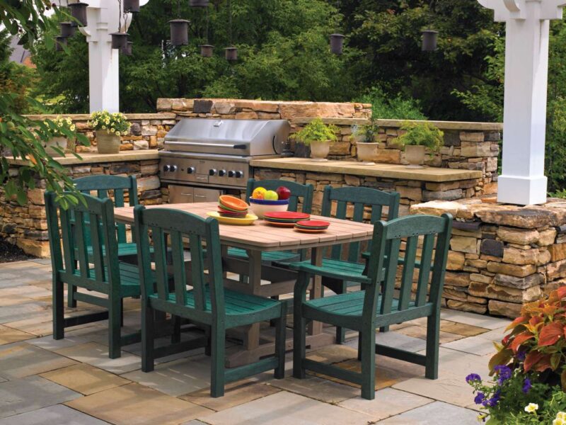 Outdoor patio setting featuring a stainless steel grill, a dining table with six green chairs, and surrounded by a stone boundary, lush plants, and an elegant fire pit.