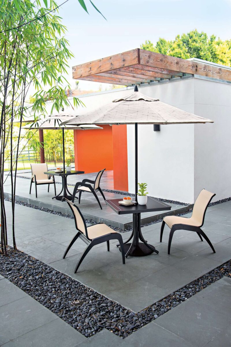 A modern outdoor patio with four chairs and a table under an umbrella, surrounded by bamboo and decorative stones, against a vibrant orange accent wall. A fire pit is inserted to enhance the ambiance.