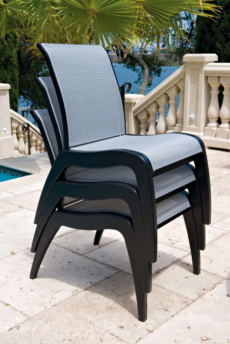A modern black outdoor armchair with a mesh back and seat, standing on a tiled patio beside a balustrade with a view of a pool and an insert fireplace in the background.