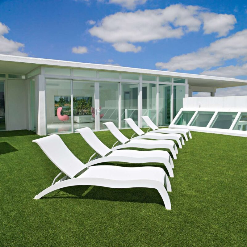 A row of white lounge chairs on a green rooftop beside a modern white building with large glass windows and an inserted fire pit under a blue sky with scattered clouds.