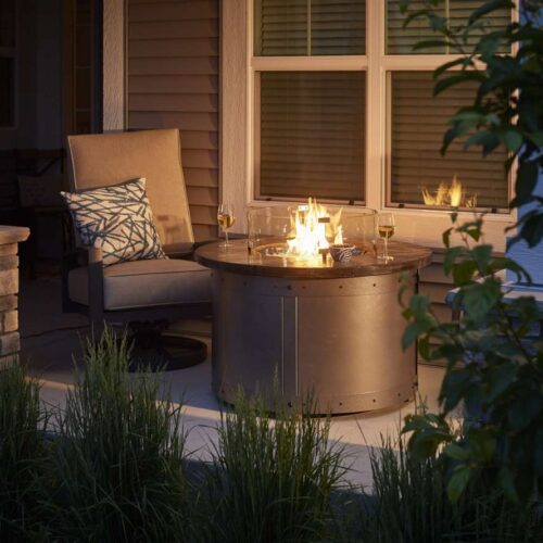 Cozy outdoor seating area by a house at dusk with a lit fire pit insert and two wine glasses, surrounded by verdant plants.