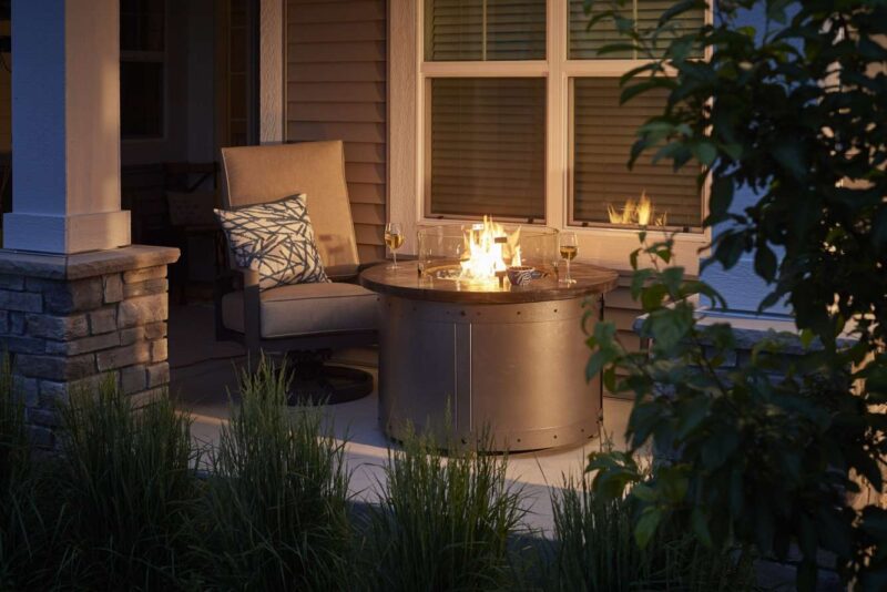 Cozy outdoor seating area by a house at dusk with a lit fire pit insert and two wine glasses, surrounded by verdant plants.