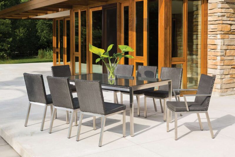 Modern patio furniture set comprising a wooden table and six woven chairs on a concrete patio beside a house with large windows, stone accents, and a fire pit.
