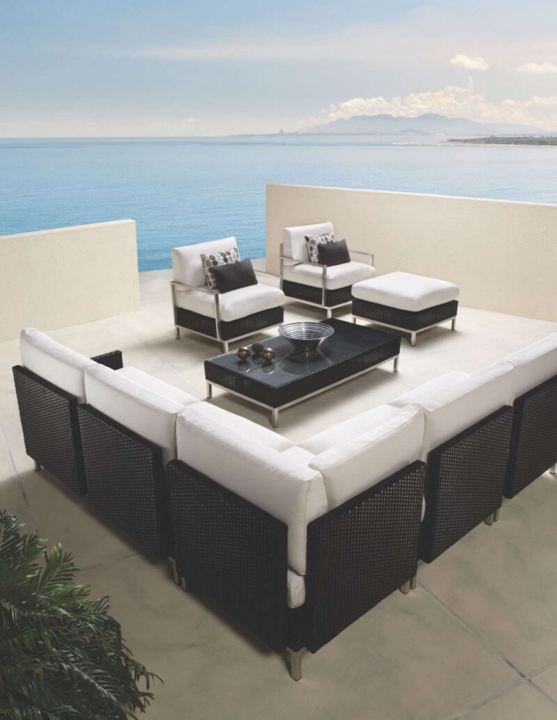 Outdoor patio with modern furniture overlooking a calm ocean. There are two armchairs, a sofa, ottomans, and a coffee table set on a tiled floor, with a fire pit nearby and