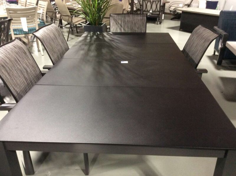 A modern black dining table set with six grey striped chairs in a furniture showroom. The table is long with a matte finish and stands prominently near a stylish fireplace, creating an inviting atmosphere amidst the various furniture