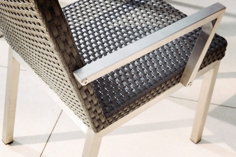 Close-up of a modern outdoor chair featuring a woven dark gray synthetic mesh seat and a sleek white metal frame, positioned near a fire pit on a tiled floor in sunlight.