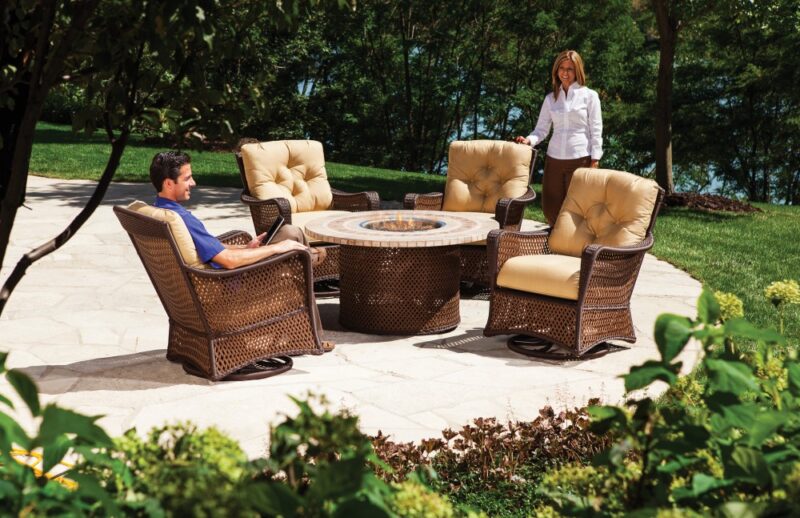 A man and a woman relax in a lush garden with outdoor wicker furniture, including plush chairs and a round table with a stone top, near a cozy fireplace, on a sunny day.