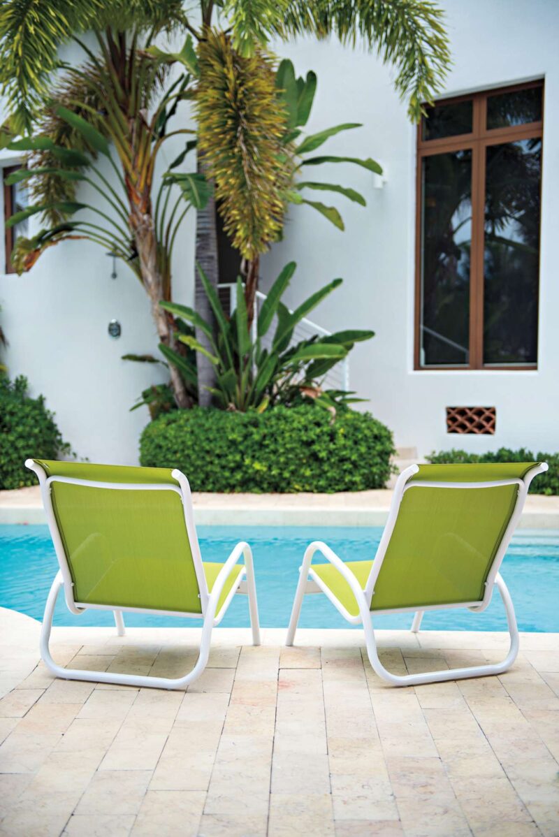 Two lime green lounge chairs on a poolside patio, facing a tranquil blue swimming pool surrounded by tropical plants and a modern house with an outdoor fire pit.