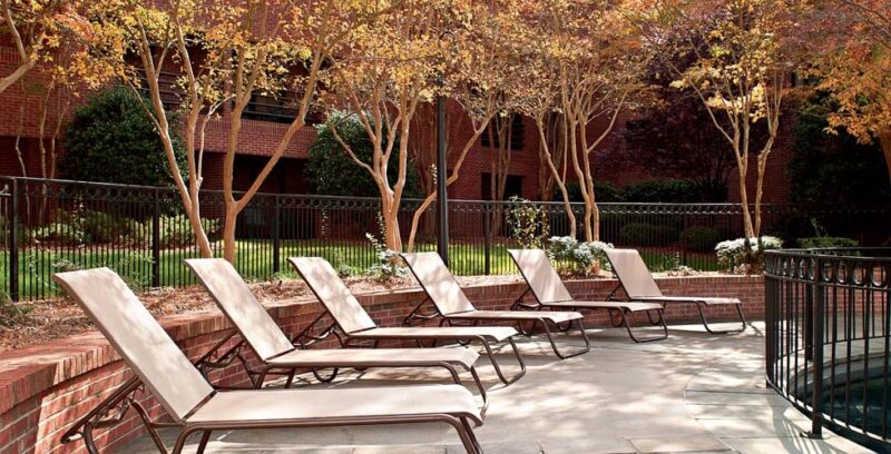 Row of beige lounge chairs on a brick patio with a fire pit, surrounded by colorful trees and a neatly trimmed hedge, with a red brick building in the background.
