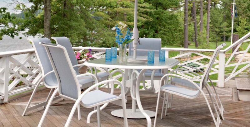 An outdoor dining setup on a wooden deck overlooking a serene lake, featuring a white table with chairs and blue table settings, surrounded by lush green trees and enhanced with an adjacent fire pit.