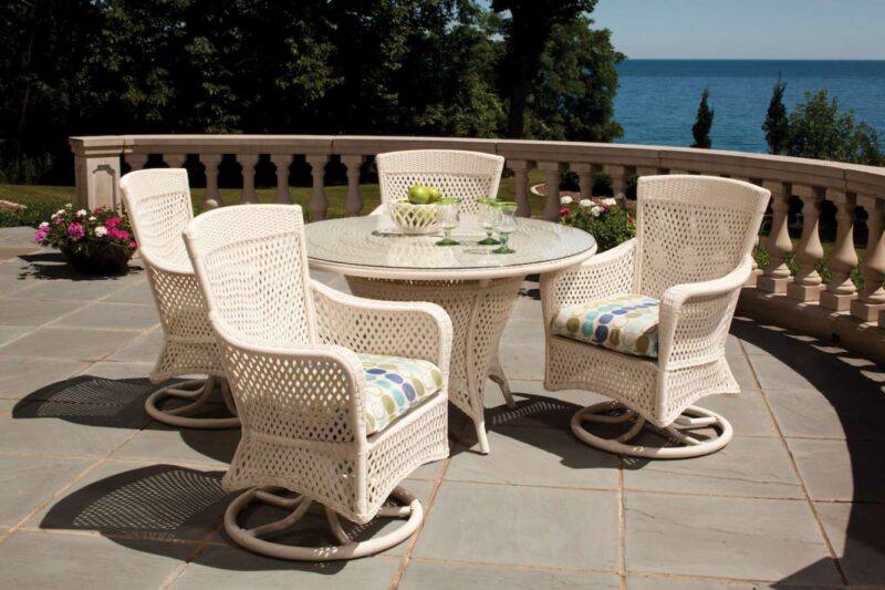 A patio overlooking the ocean features a table and four chairs with comfortable cushions, set on a flagstone floor, surrounded by lush greenery and a fire pit on a sunny day.