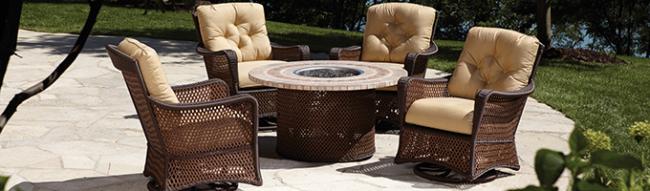 A luxury outdoor seating area featuring a circular table with a built-in fireplace, surrounded by wicker chairs with plush beige cushions, set against a background of lush greenery.