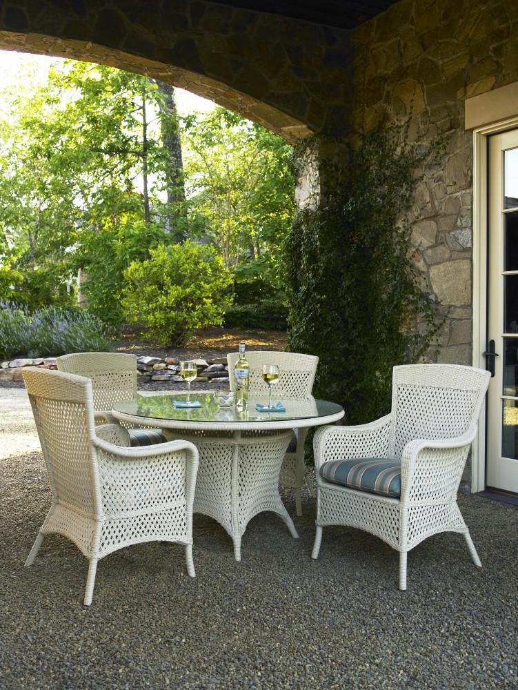 An outdoor dining area under a stone archway with a round table, four white wicker chairs, and a view of a lush garden and stone wall. Two wine glasses and a bottle are on the