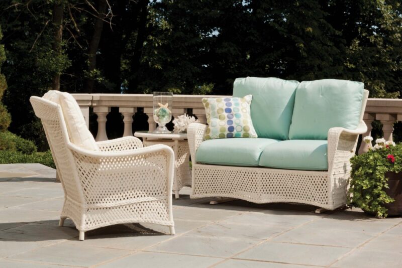 White wicker patio furniture with light blue cushions on a stone terrace surrounded by lush greenery and a cozy fireplace insert in a sunny outdoor setting.
